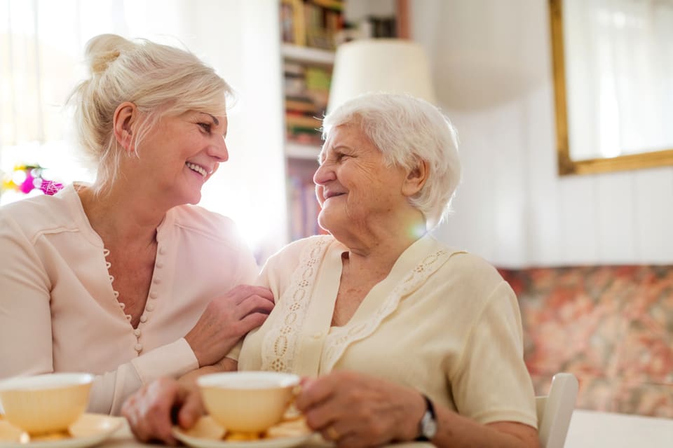 A senior and middle aged woman laugh while sipping from tea cups.